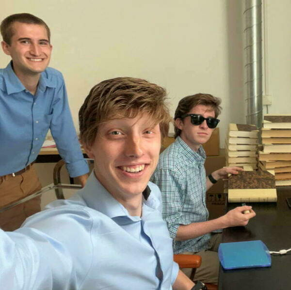 Three undergraduate men in coordinating blue button-down shirts pause from their work in the archives for a selfie.