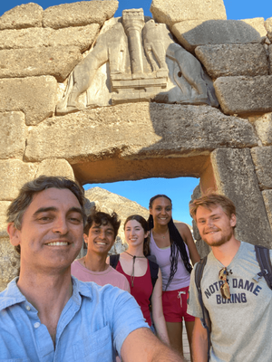 Five people smile for a selfie beneath the Lion Gate at Mycenae, Greece.  The ancient gate's stone relief depicts two lions flanking a central column.
