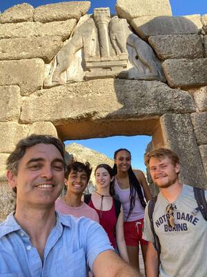Five people smile for a selfie beneath the Lion Gate at Mycenae, Greece.  The ancient stone gate features a carved relief of two lions flanking a column.  A hillside is visible through the gate.