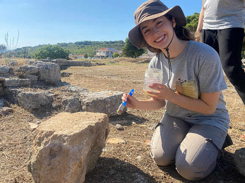 A smiling student wearing a wide-brimmed hat and gray shirt kneels at an archaeological site.  She holds a small brush and a jar of liquid, applying it to a large, light-brown stone.  Partial ruins and a building are visible in the background.