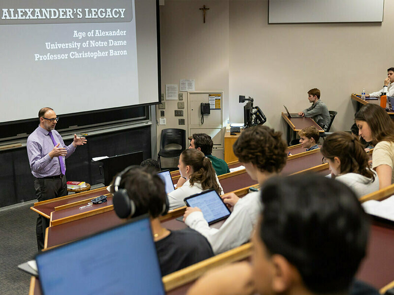 Professor Christopher Baron, wearing a purple shirt and tie, gestures while lecturing to students in a University of Notre Dame classroom.  The projection screen displays the lecture title, "Alexander's Legacy," along with Baron's name and affiliation.  Students sit at tiered desks, some using laptops and tablets.