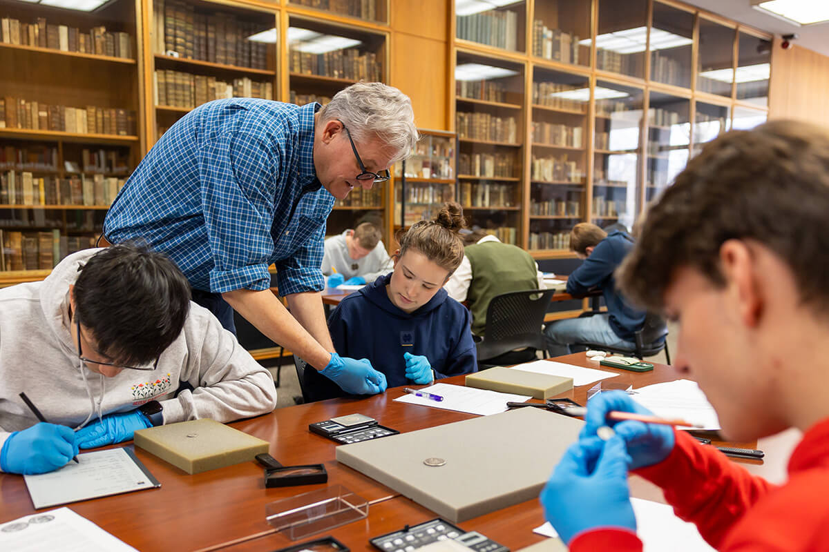 A professor wearing a blue plaid shirt and blue gloves leans over a table, assisting students examining small artifacts.  Students wear blue gloves and work with tools like magnifying glasses and small scales in a room with floor-to-ceiling bookshelves.