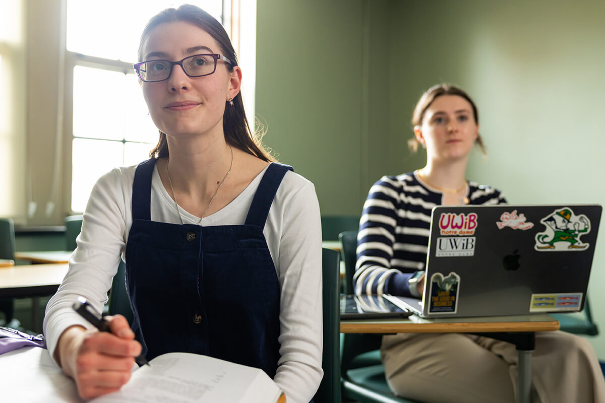 A female student wearing glasses and a navy blue jumper looks towards the front of a classroom, holding a pen over her open notebook.  Another student in a striped shirt works on a laptop in the background.