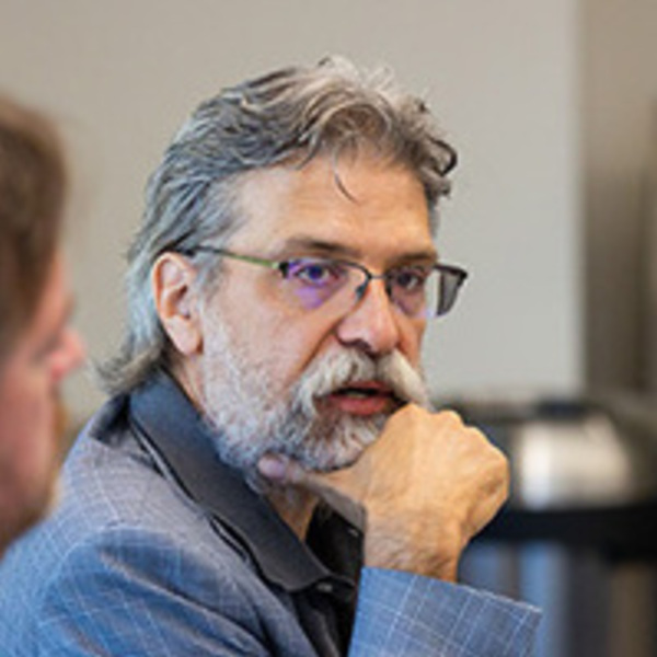 Bearded man with gray hair and glasses, wearing a light blue plaid jacket, intently listens with hand to chin.
