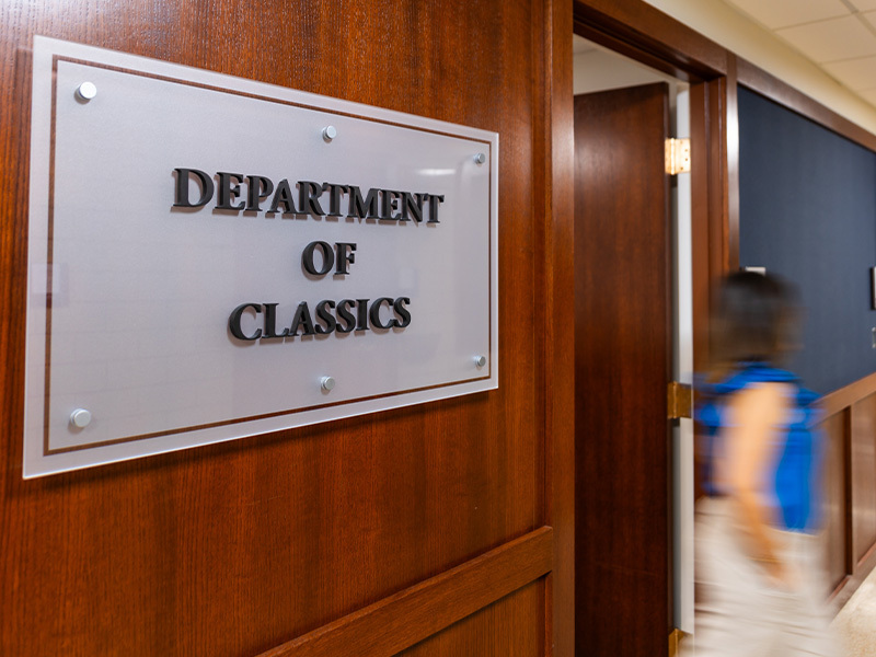 Frosted glass sign reading "Department of Classics" on a varnished wooden office door. A blurred figure in a blue shirt walks past in the hallway.