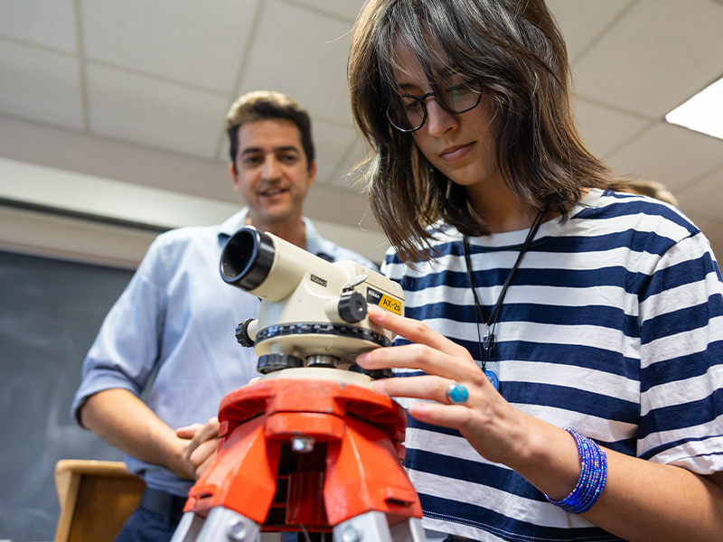 A student wearing a navy and white striped shirt adjusts a theodolite on a red tripod while a professor looks on.
