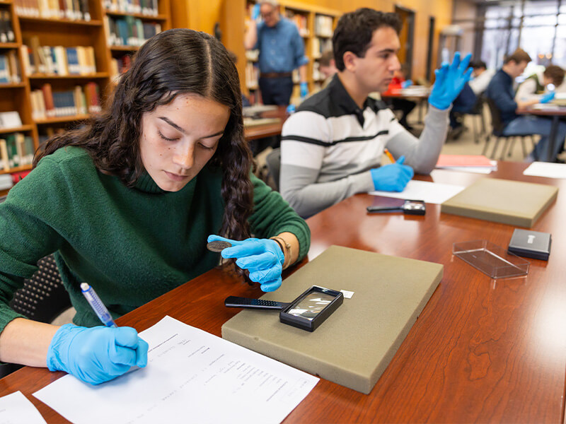 A student wearing a green sweater and blue gloves examines a small artifact while taking notes in a library. Another student and a blurred figure are visible in the background.