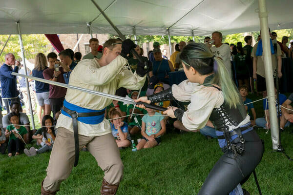Two costumed performers duel with swords on green grass under a white tent. A person in a cream tunic and blue sash lunges at another in a black corset and dark pants, holding a sword defensively. An attentive audience of adults and children watches.