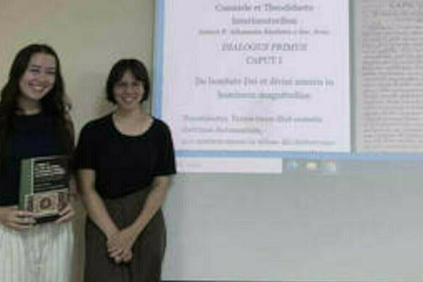 Two smiling women stand before a presentation screen showing academic text. One woman wears a dark top and striped pants, holding a green book. The other wears a dark top.