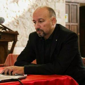Man with a short beard and receding hairline in a black suit jacket and dark shirt sits at a red draped table, typing on a laptop. A microphone and a wooden lectern are visible in the blurry background.