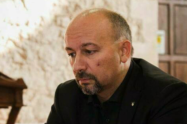 A bald man with a dark beard, wearing a black suit and shirt, sits at a red-covered table, looking down intently. His hands rest on papers in front of him. A microphone and wooden furniture are in the background.