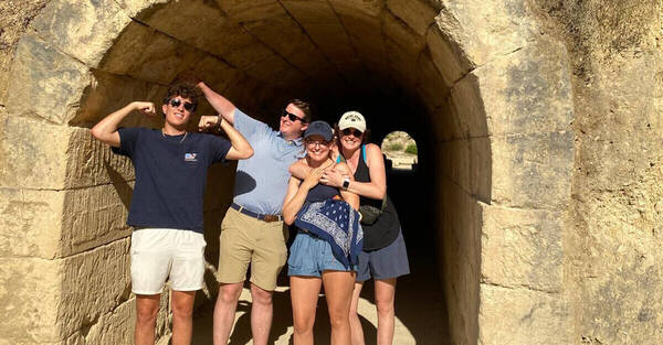Four smiling young adults stand at the rough-hewn stone entrance of an ancient-looking tunnel. A man on the left flexes, while two women embrace, and a second man has his arm around one. The tunnel extends to a bright opening revealing arid hills.