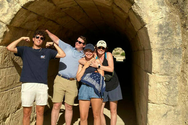 Four smiling young adults stand at the rough-hewn stone entrance of an ancient-looking tunnel. A man on the left flexes, while two women embrace, and a second man has his arm around one. The tunnel extends to a bright opening revealing arid hills.
