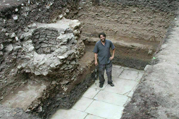A smiling man with dark hair and beard, wearing a striped shirt, stands in an archaeological trench. He holds a measuring stick next to a round stone well-like structure and light gray paved floor. The trench walls show layers of dark earth.