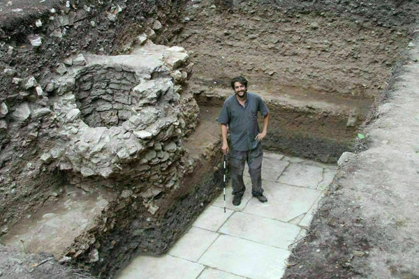 A smiling man with dark hair and beard, wearing a striped shirt, stands in an archaeological trench. He holds a measuring stick next to a round stone well-like structure and light gray paved floor. The trench walls show layers of dark earth.