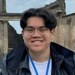 Young man with dark hair, gold glasses, and a black jacket smiles broadly, wearing blue earbuds and lanyard, in front of ancient stone ruins.