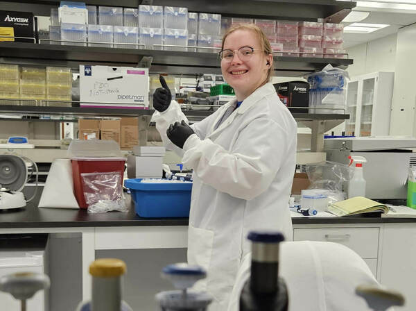 A smiling young woman in a white lab coat and black gloves gives a thumbs-up in a science laboratory. Shelves filled with lab supplies and various equipment like pipettes and a centrifuge are visible around her.