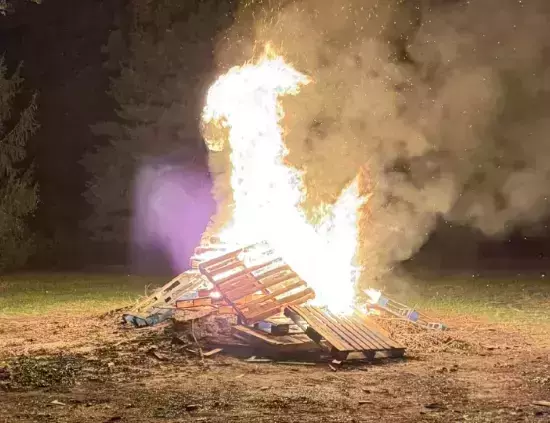 A large bonfire, built from wooden pallets, burns intensely at night. Bright white and orange flames with sparks rise above dense smoke. Trees are visible in the dark background.