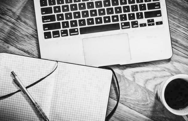 Black and white photo of a laptop keyboard, a gridded notebook with a pen, and a cup of coffee on a wooden desk.