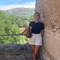A smiling East Asian woman in a black t-shirt and striped light shorts stands by an old brick wall with a diagonal pattern. Behind her is a green valley with a town on a distant hillside under a blue sky.