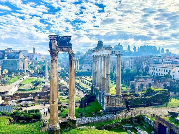 Elevated view of the Roman Forum: weathered columns, green grounds, walking paths, and distant buildings under a blue, cloudy sky.