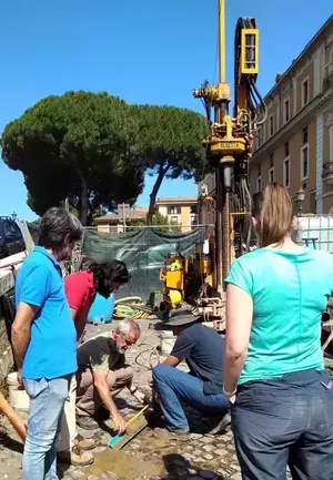 Five people examine a soil sample from an excavation pit on a cobblestone street. A large yellow drilling rig is in the background.