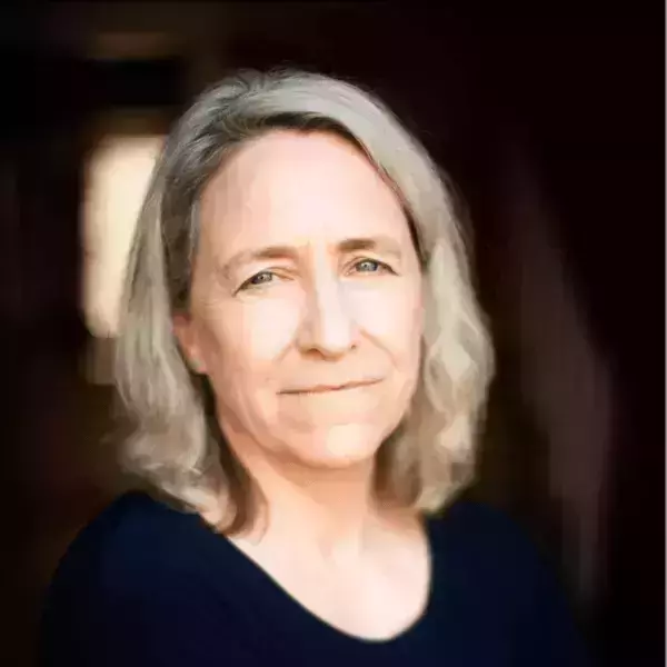 A woman with wavy, shoulder-length grey hair, blue eyes, and a slight smile wears a dark blue shirt.