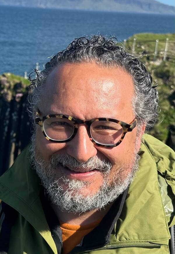 Smiling man with gray curly hair, beard, tortoise-shell glasses, green jacket, near ocean cliffs.