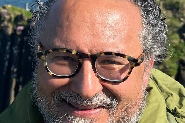 Smiling man with gray curly hair, beard, tortoise-shell glasses, green jacket, near ocean cliffs.
