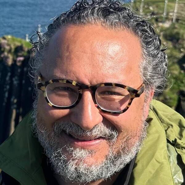 Smiling man with gray curly hair, beard, tortoise-shell glasses, green jacket, near ocean cliffs.