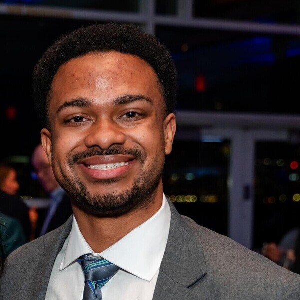 Young Black man with a beard, wearing a grey suit and striped tie, smiles broadly.
