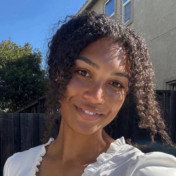 Smiling woman with dark curly hair wears a white ruffled top in bright outdoor sunlight.