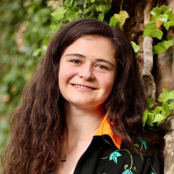 Smiling woman with long brown hair, black jacket with orange collar and green floral embroidery, wearing a cross necklace, by ivy.